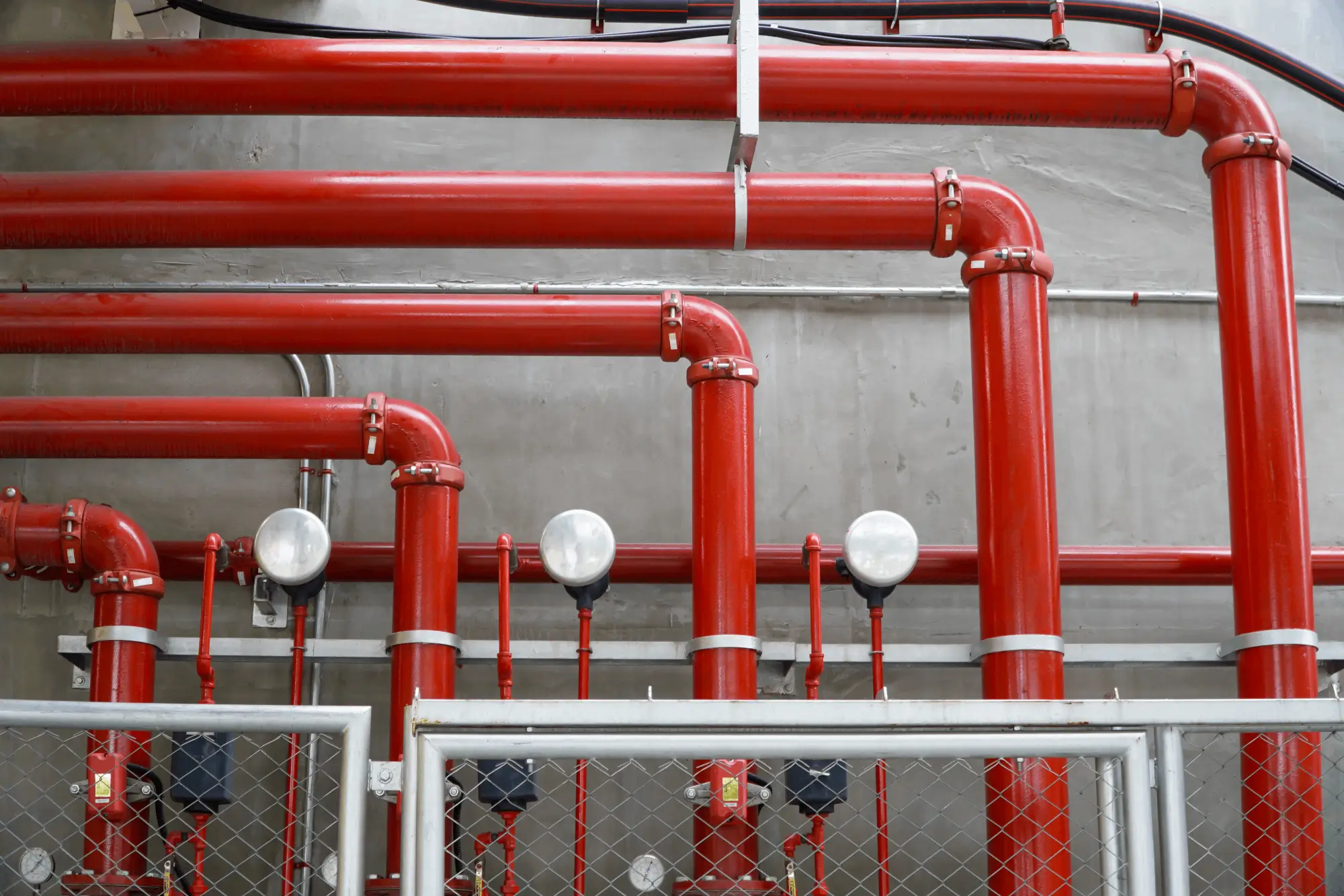 Ein komplexes Netzwerk aus roten Rohrleitungen einer Sprinkleranlage vor einer grauen Betonwand, mit Druckventilen und Messgeräten.