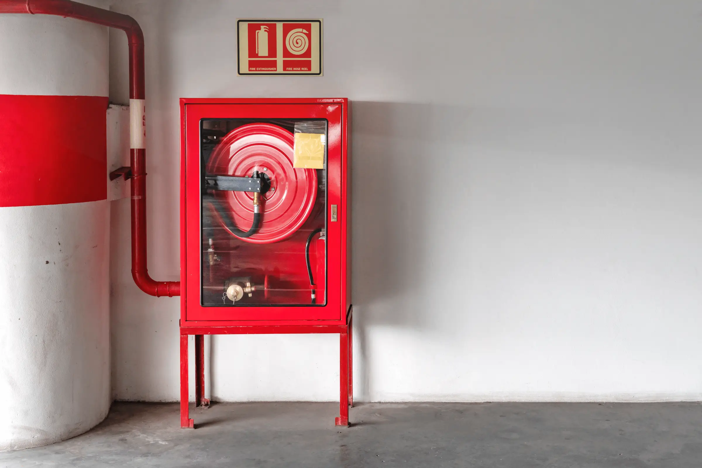 Ein Wandhydrant mit einem roten Gehäuse und einer darin aufgerollten Feuerwehrschlauchrolle, montiert an einer weißen Wand mit einem Brandschutzsymbol darüber.